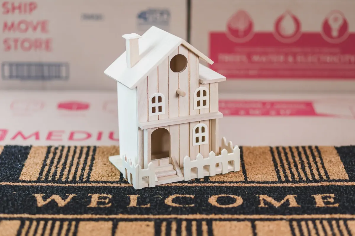 A small wooden house model sits on a welcome mat among moving boxes, symbolizing a new home.