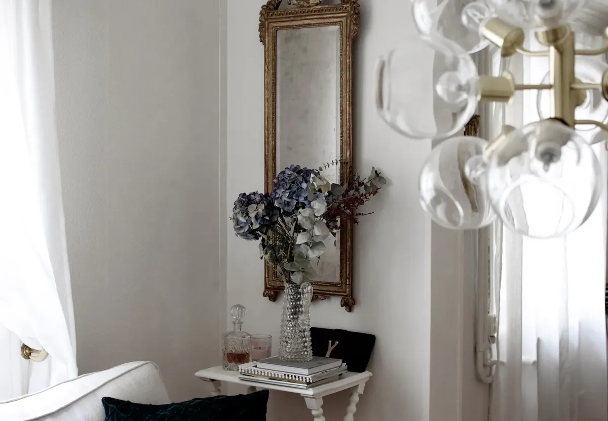 Interior of elegant room with table with vase with bouquet of various flowers near mirror on wall and curtains next to sofa with pillows under lamp